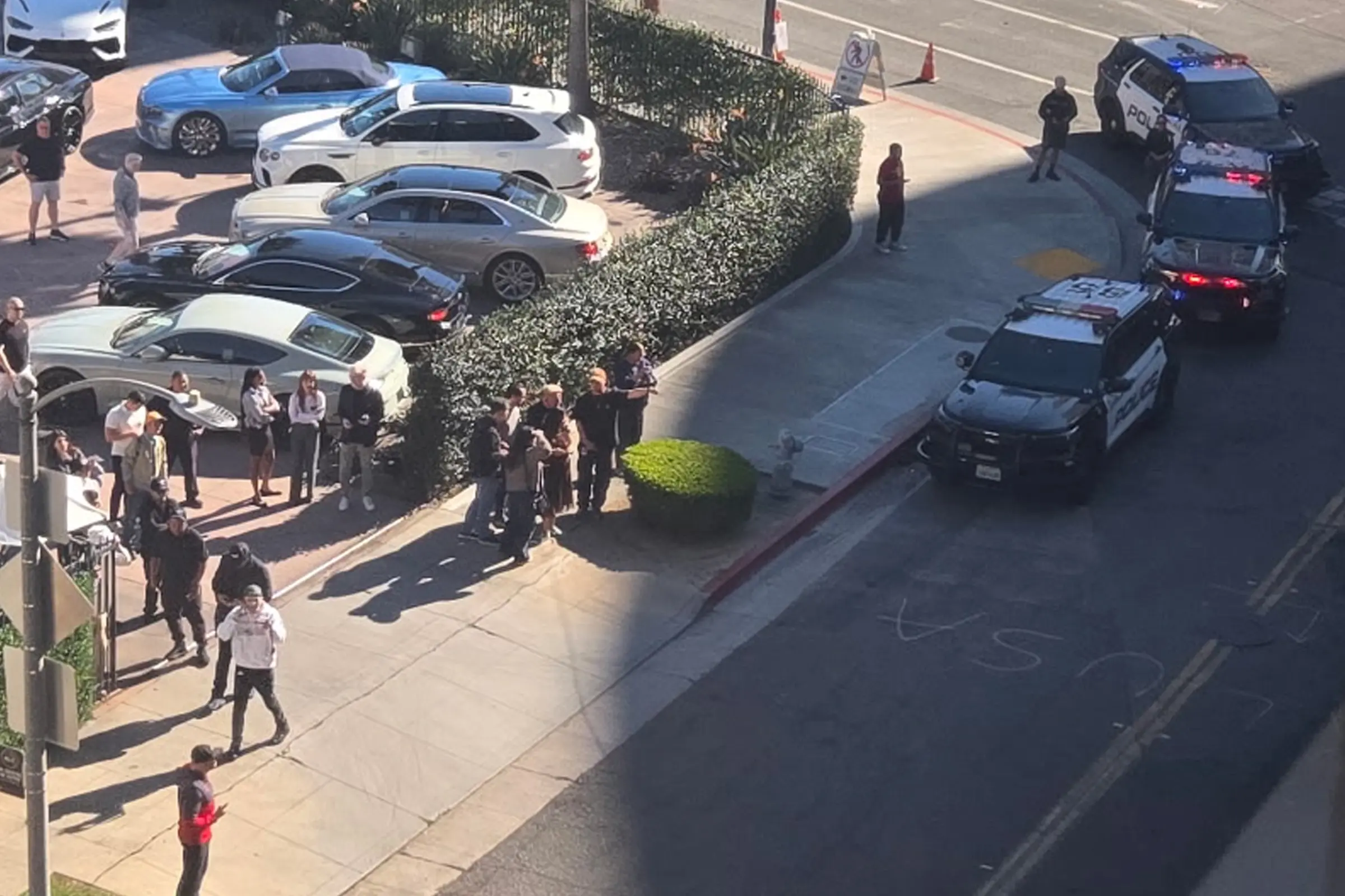 Beverly Hills police secure Wilshire Boulevard near San Vicente Boulevard after a serious mid-day assault, drawing the attention of nearby pedestrians and office workers overlooking the scene.
