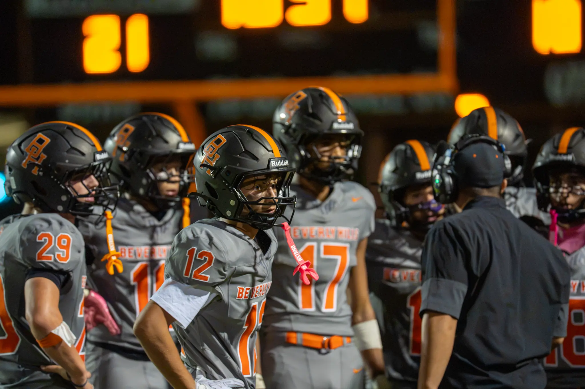 Beverly Hills football players gather around a coach wearing a headset on the sideline.