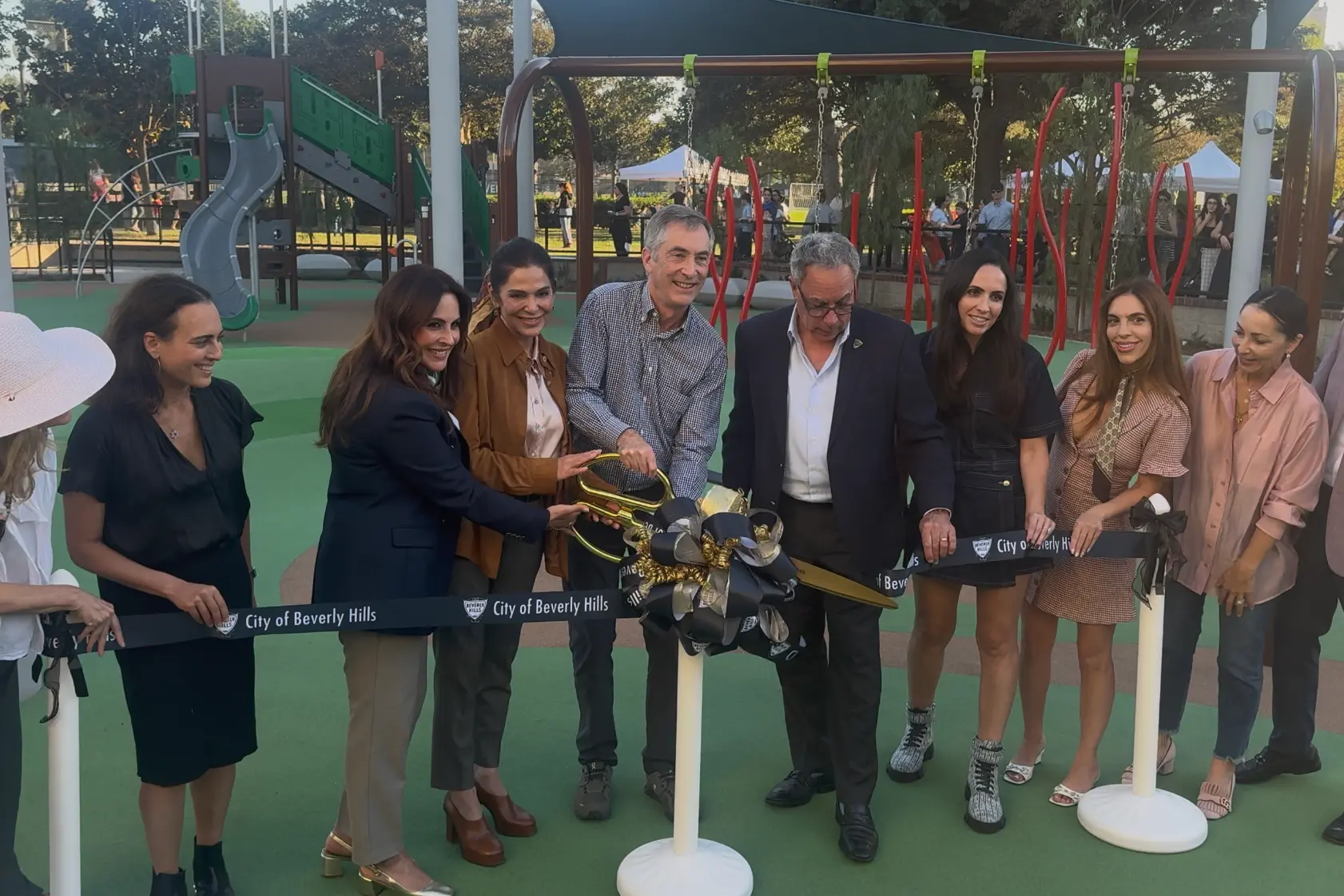 Ribbon-cutting participants hold oversized gold scissors and a City of Beverly Hills ribbon in front of playground equipment at La Cienega Park.