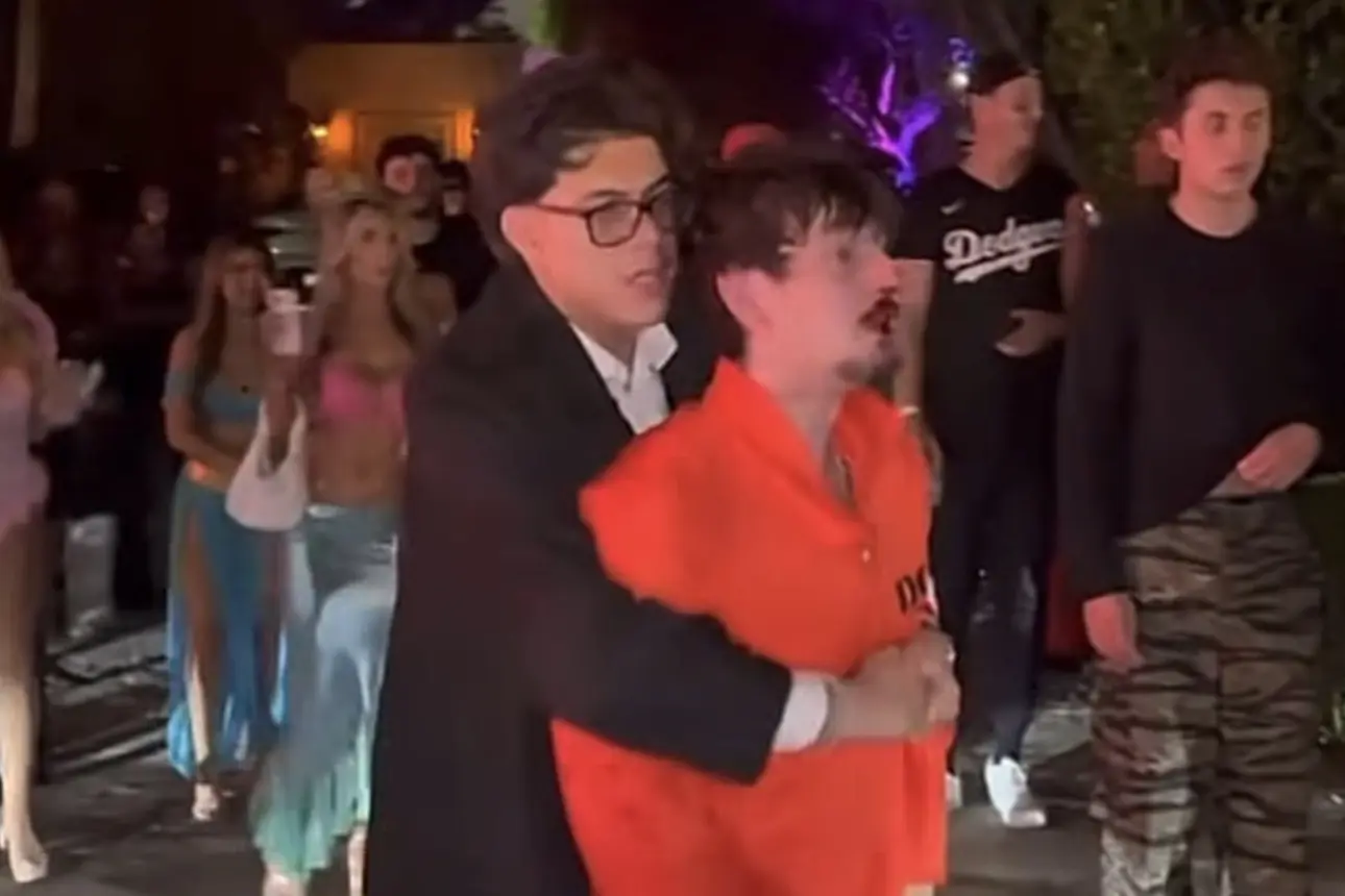 Bryce Hall in an orange costume with blood visible around his nose, accompanied by a man in dark clothing as partygoers stand nearby at night.