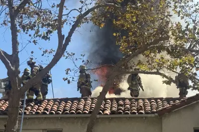 Firefighters work on the roof of an apartment complex in the 200 block of South Rexford Drive in Beverly Hills as flames and heavy smoke rise behind them.