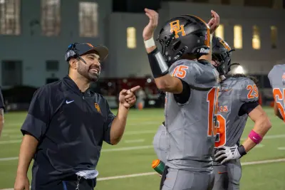 A smiling Beverly Hills coach stands beside football players, including a player raising his hands.